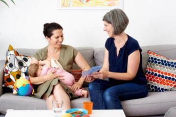 Eine Frau stillt ihr Kind während eine zweite Frau ihr Broschüren zeigt