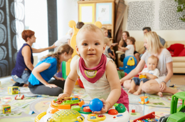 Baby beim Spielen. Im Hintergrund sind Eltern und Babys in einer Gruppe beim spielen und sprechen zu sehen.